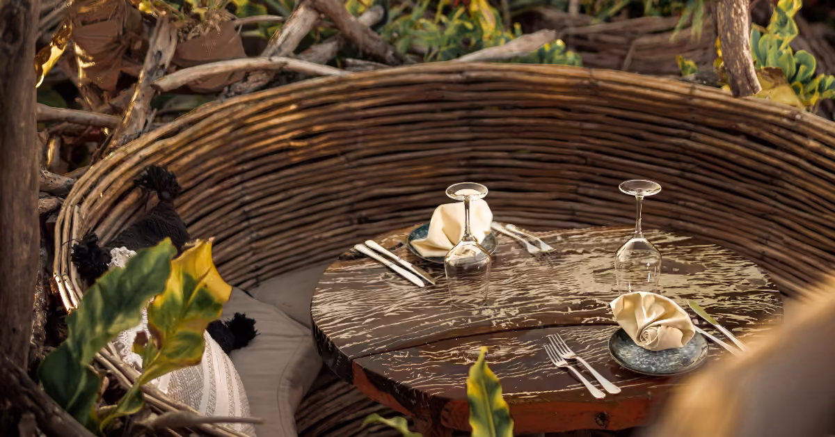 A round wooden table set for two with napkins and glassware, surrounded by branches and greenery for a cozy dining nook.