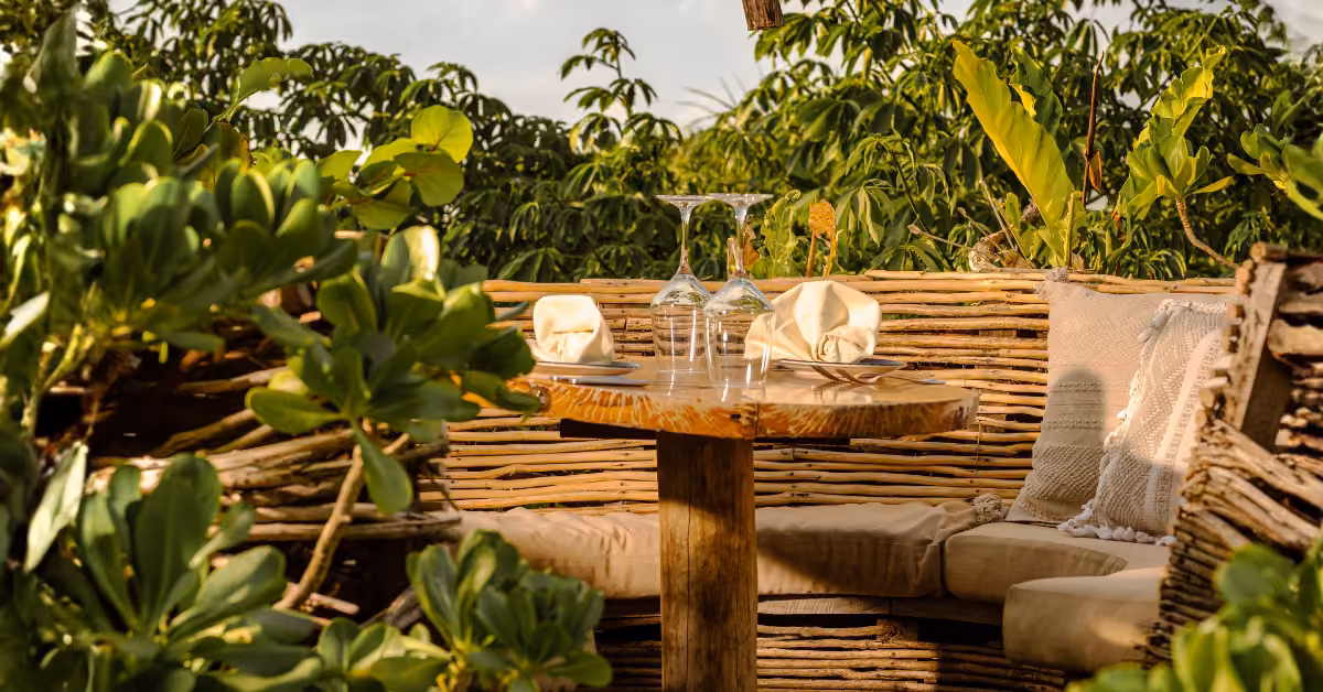 A cozy outdoor table for two with cushioned benches, greenery, and woven fencing, set with plates and glasses in sunlight.