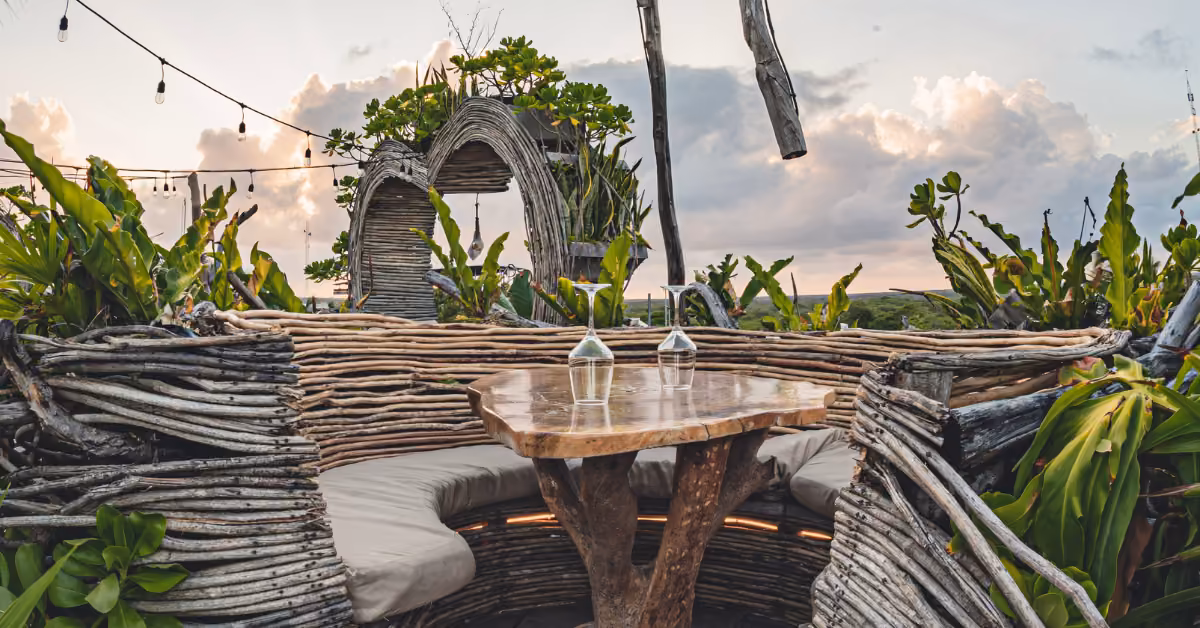 Karma Rooftop’s cozy outdoor spot has a wooden table, wicker benches, greenery, and string lights under a sunset sky.