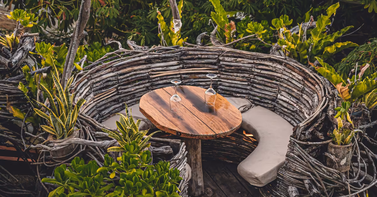 A round wooden table with two upside-down wine glasses sits in a rustic branch bench, surrounded by lush greenery outdoors.