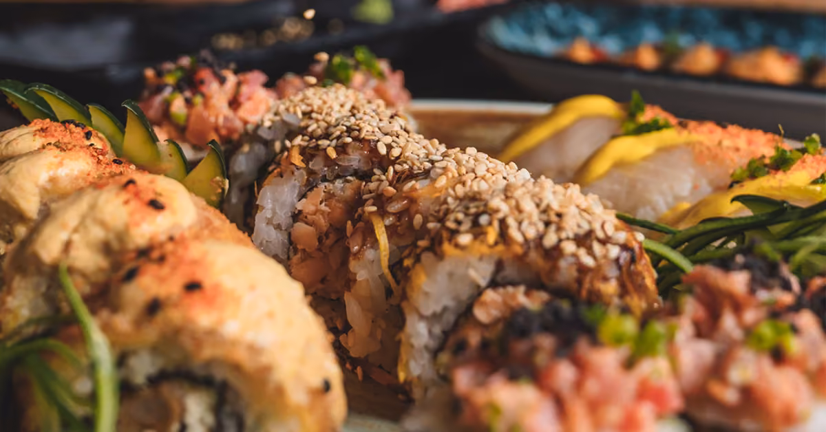 Assorted sushi rolls topped with sesame seeds, sauces, and garnishes on a plate, with sliced avocado in the background.