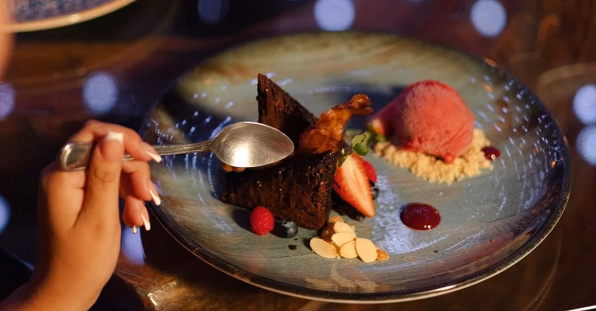 A person holds a spoon over chocolate cake, berries, almond slices, and pink ice cream, an elegant dessert at Karma Rooftop.