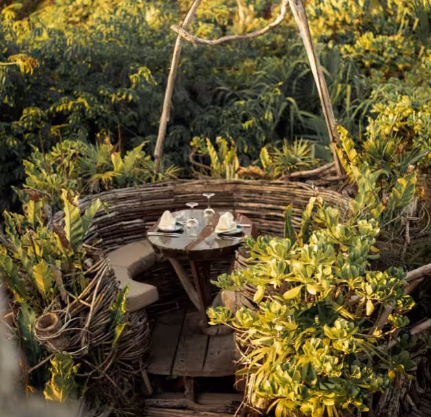 An outdoor table for two with plates and wine glasses sits in a wooden nook on a rooftop, surrounded by lush greenery.