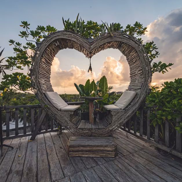 A heart-shaped frame with two seats faces a small table on a rooftop deck, surrounded by greenery at sunset.