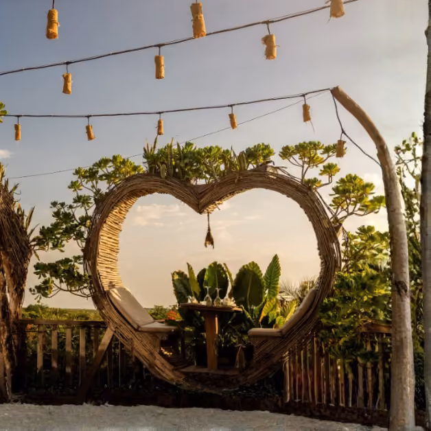 Heart-shaped wicker chairs on a wooden deck with plants, string lights, and sky views create a romantic rooftop setting.