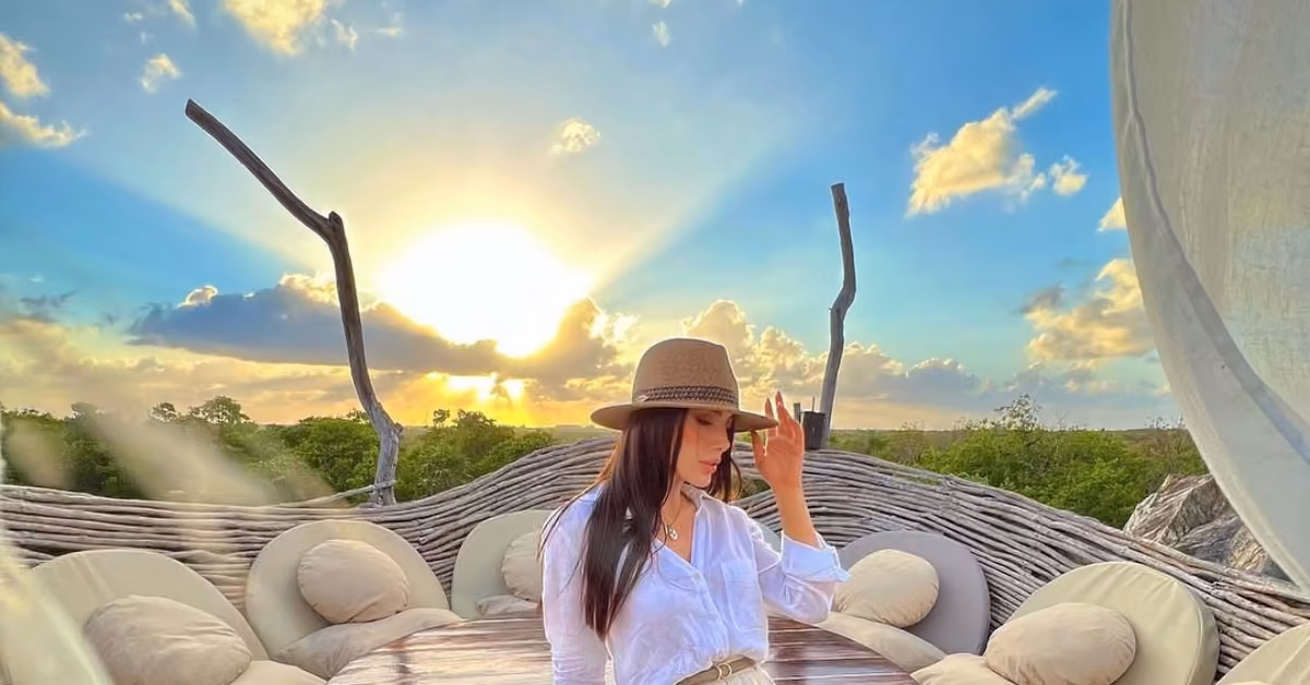 A woman in a white shirt and brown hat sits on a round outdoor sofa at sunset, framed by woven branches under a blue sky.