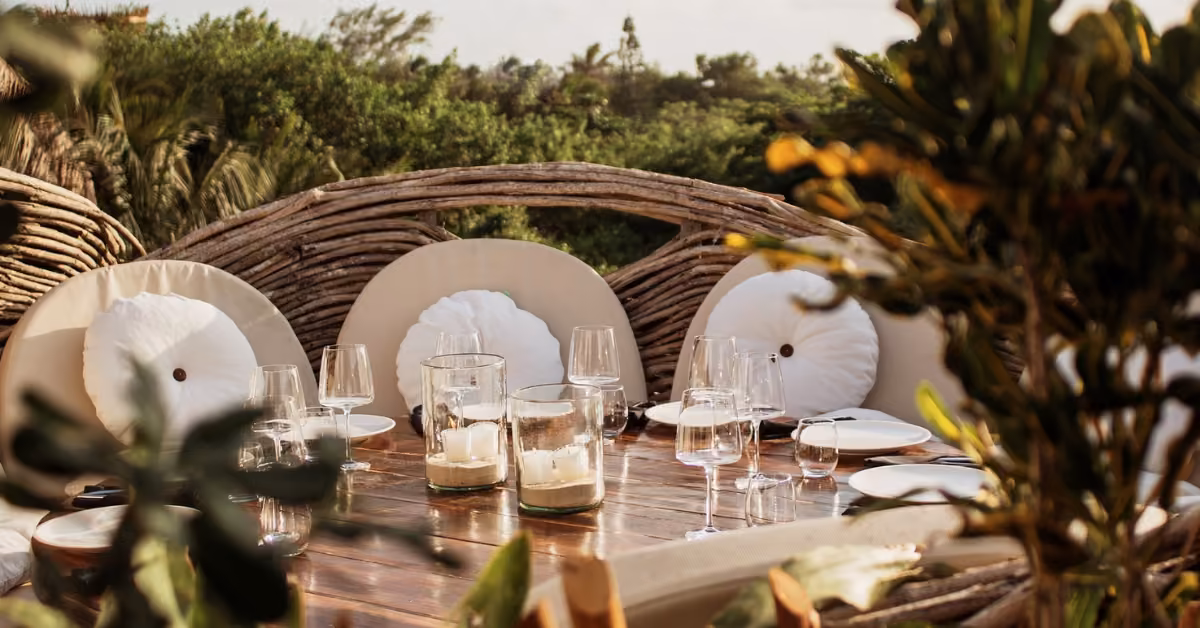 A rustic outdoor dining table with plates, glasses, candles, curved wooden chairs, and lush greenery creates a relaxed setting.