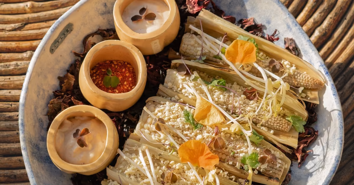 Tamales in a bowl, topped with baby corn, edible flowers, and seeds, served with creamy sauces on a rustic woven surface.
