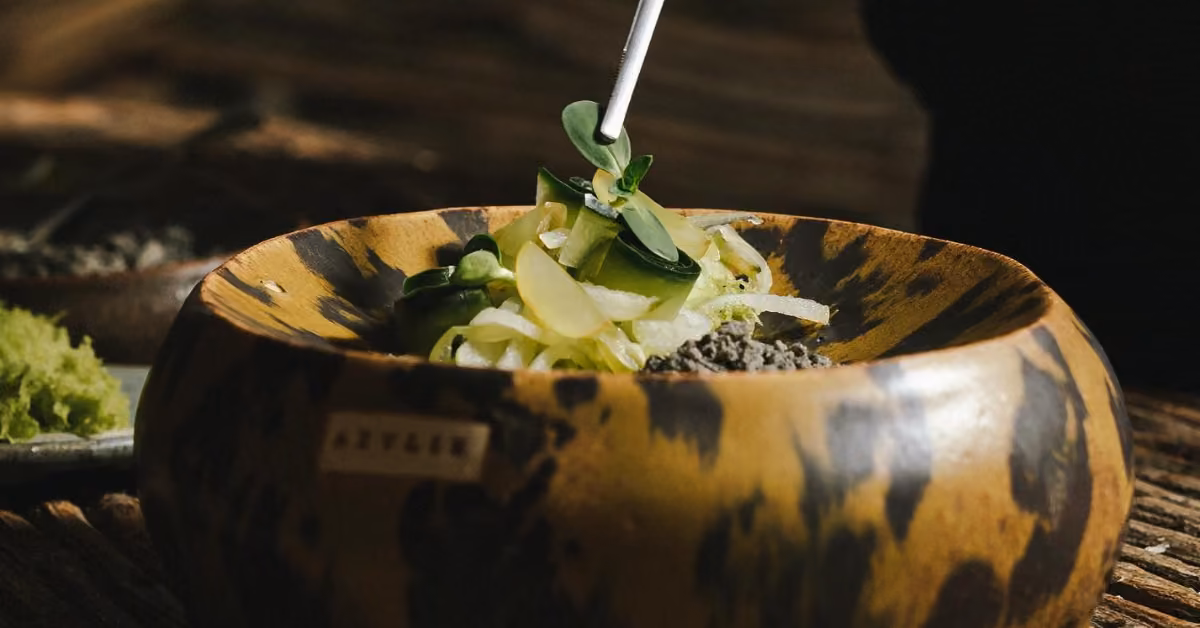 Wooden bowl with a mottled pattern, filled with fresh greens, sliced veggies, and seeds. A fork rests inside, ready to eat.