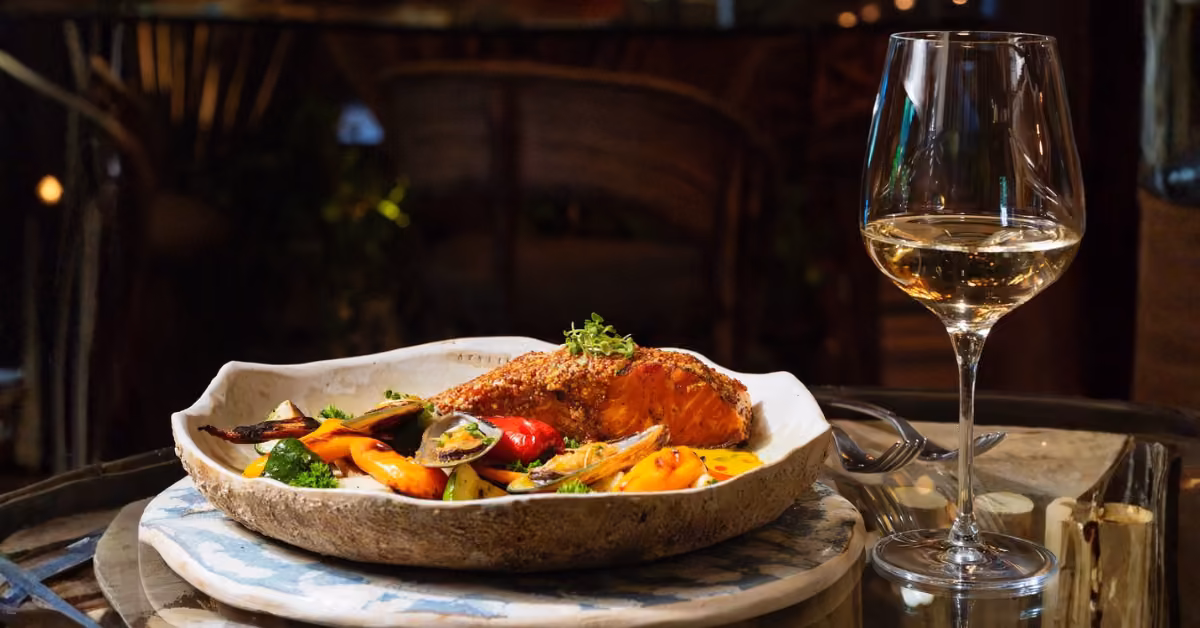 Grilled salmon with mixed veggies and a glass of white wine sit on a rustic table, with fork and knife set beside the plate.