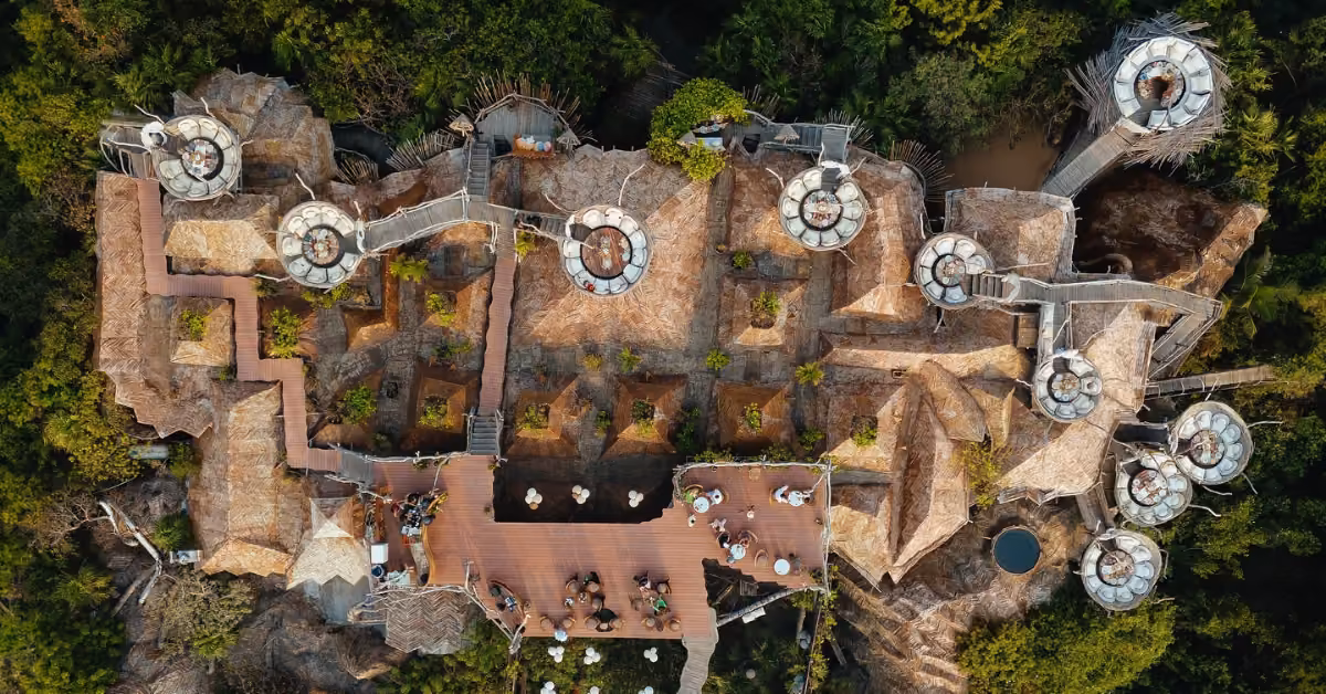 Aerial view of a resort with thatched roofs, circular towers, wooden walkways, outdoor dining areas, and lush green trees.