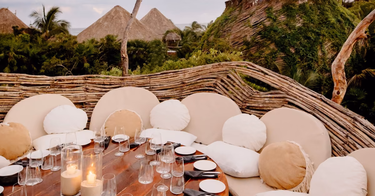 A round dining table with plates, glasses, and candles is surrounded by cushioned seats, greenery, and thatched-roof huts.
