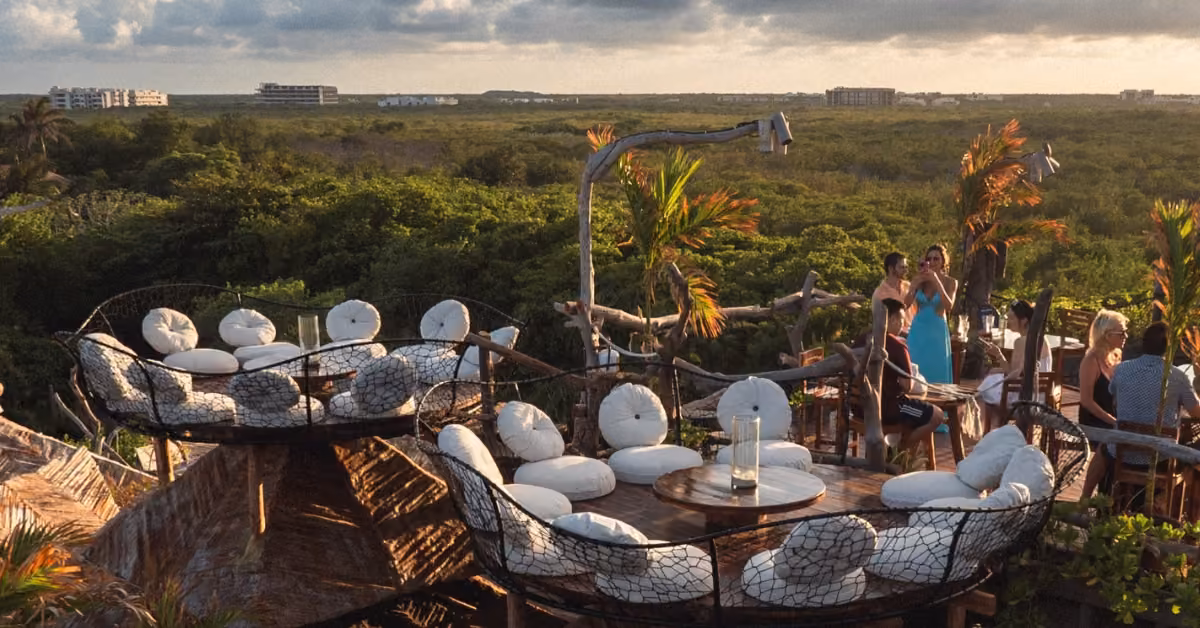 Kin-Toh rooftop bar with circular seating, cushioned chairs, people socializing, lush greenery, and a sunset sky.