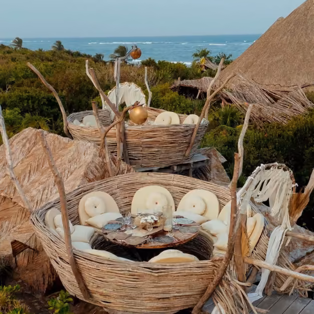 Wicker nest-style seats with beige cushions and a table, set amid greenery and ocean views, create a cozy tropical dining spot.