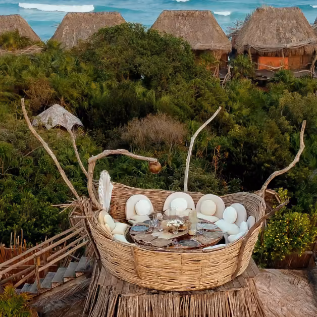 Woven wood nest seating with a round table and white cushions overlooks greenery, thatch huts, and the ocean on a rooftop.