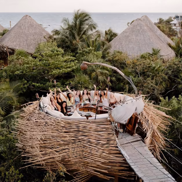 People enjoy a circular outdoor dining area on a rooftop surrounded by greenery, huts, and ocean views, some raising their hands.