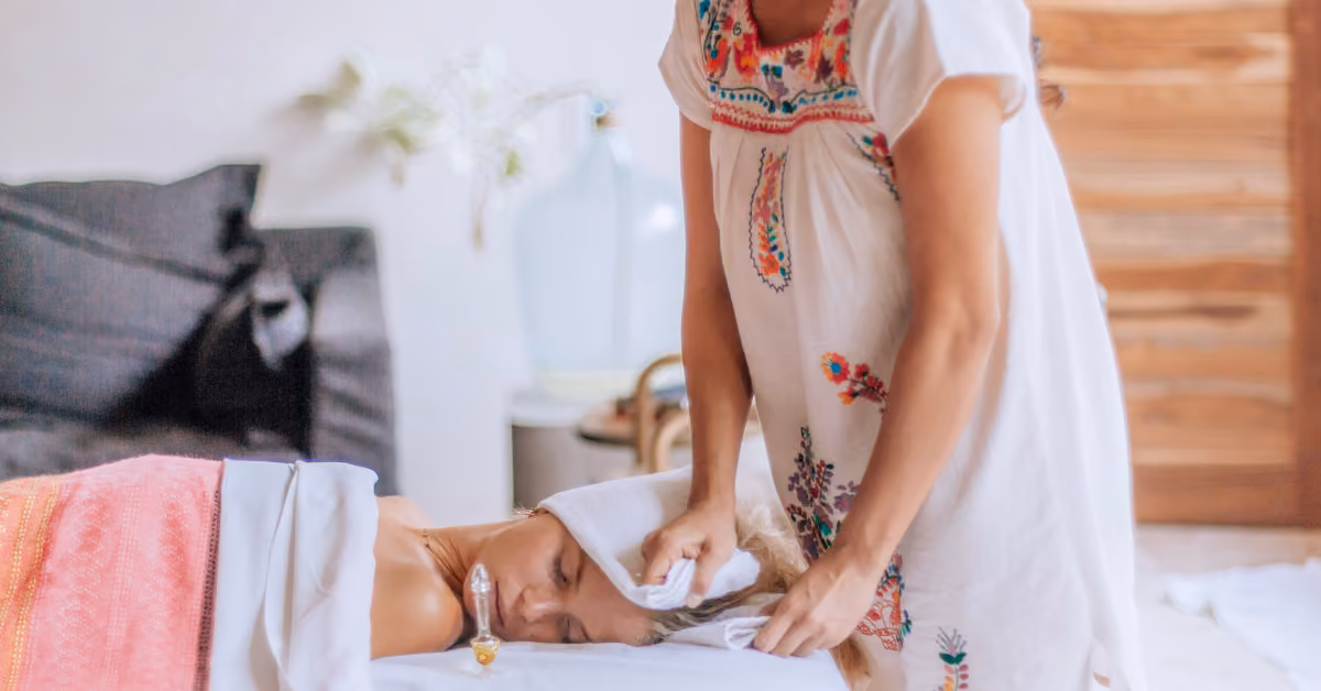 A person relaxes on a massage table with eyes closed as someone gently wipes their face in a calm spa environment.