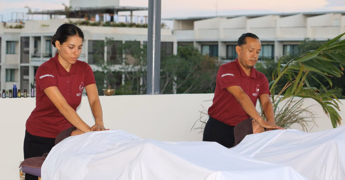Massage therapists in maroon shirts work on clients with white sheets outdoors, surrounded by modern buildings and plants.