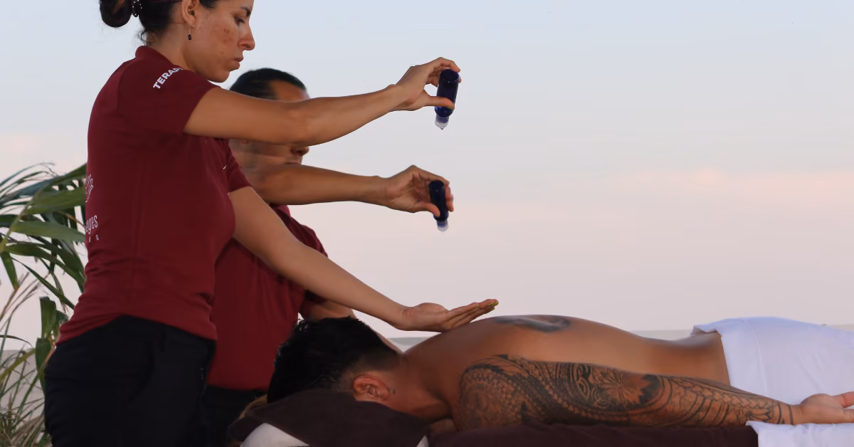 Two therapists in maroon shirts pour oil onto their hands, preparing to massage a man lying face down on an outdoor table.