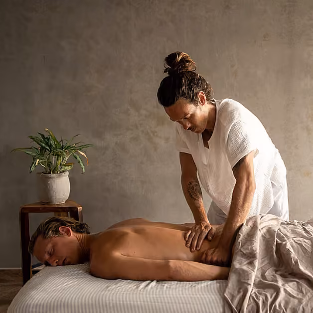 A person gets a back massage in a softly lit, neutral room. A small plant rests on a table beside the massage bed.
