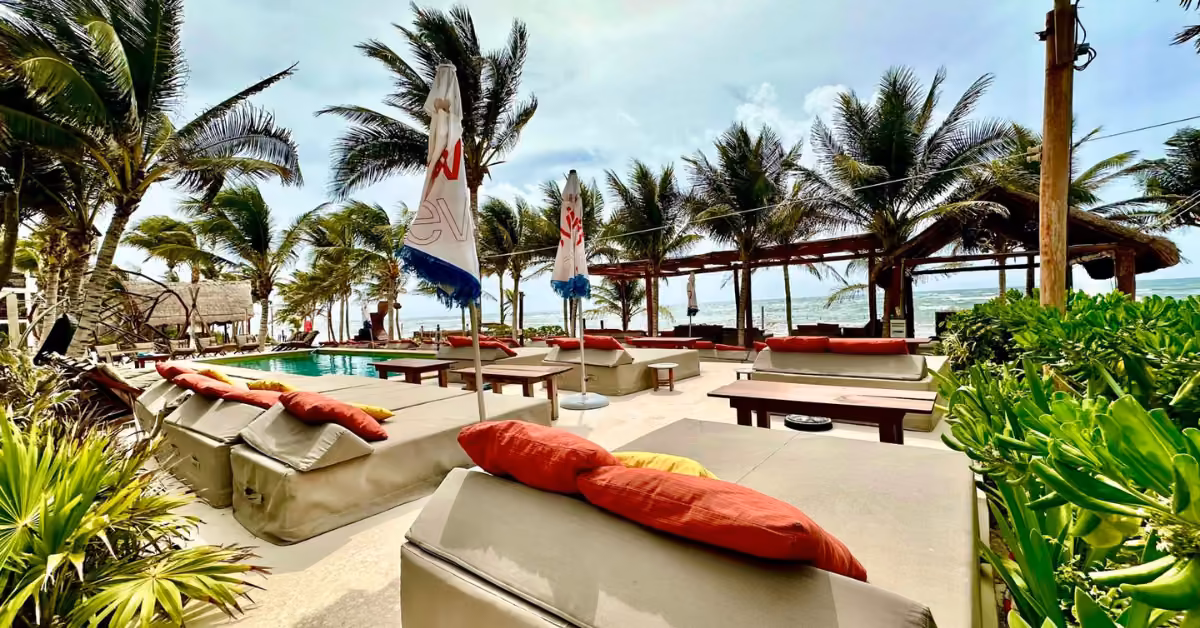 Mia Beach Club’s outdoor lounge has cushioned beds, bright pillows, palm trees, umbrellas, and a pool facing the ocean.