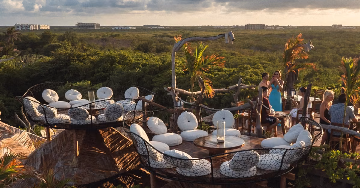 Rooftop bar with round seating, white cushions, and wood tables among greenery overlooks the coast. Guests relax and enjoy the view.