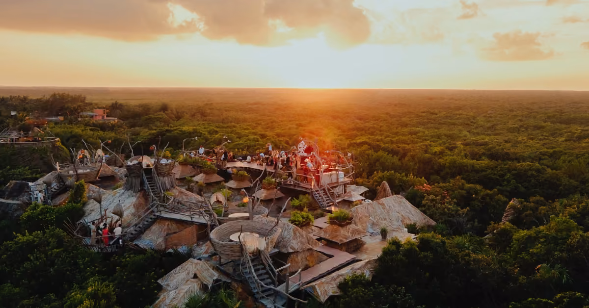 An elevated wooden treehouse overlooks lush jungle at sunset, as people enjoy golden sky views from its rooftop terraces.