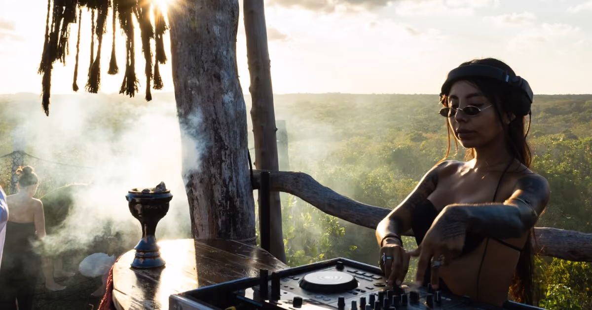 A woman in headphones and sunglasses DJs on a rustic rooftop at sunset, smoke swirling as sunlight filters through the trees.
