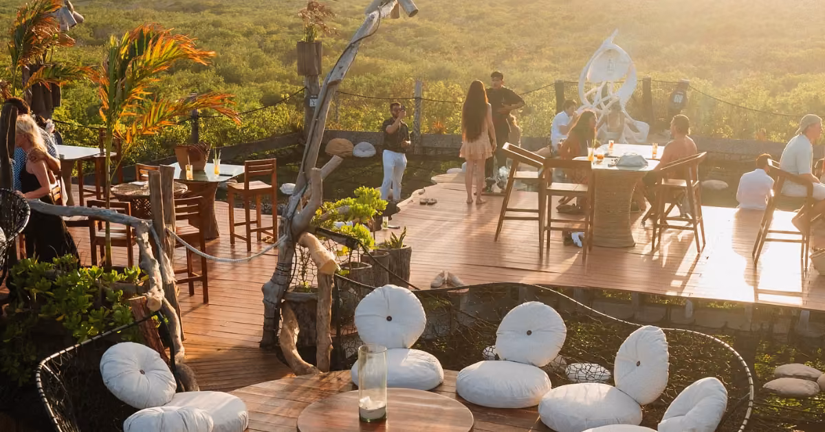 Sunny rooftop deck with wicker chairs, wooden tables, people socializing, and greenery in a relaxed, inviting atmosphere.