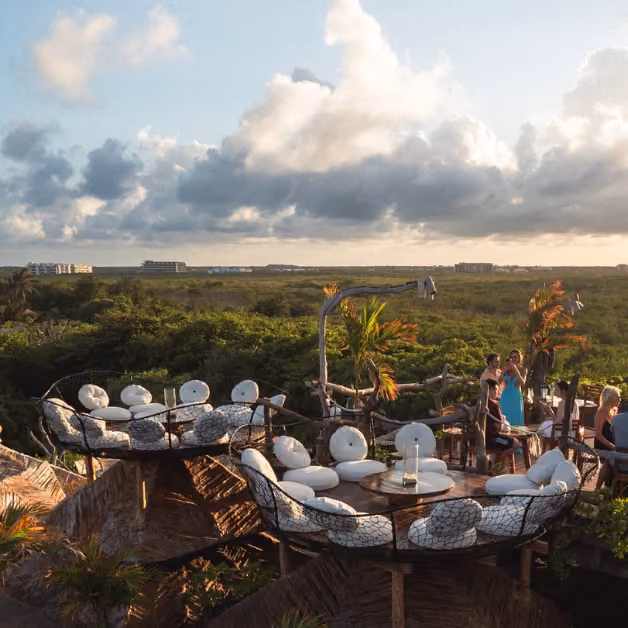 Rooftop lounge with round tables and chairs overlooks greenery at sunset; people relax and enjoy the scenic view.
