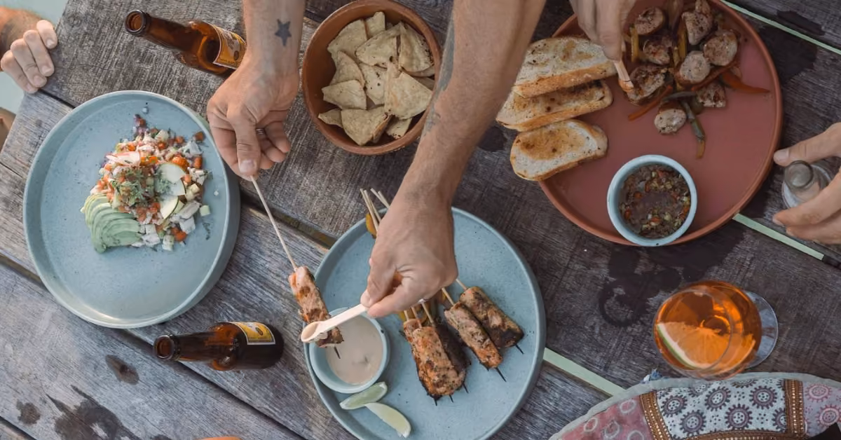 People share an outdoor meal at Neek Lagoon Club: grilled skewers, avocado salad, beer, and hands reaching for food.