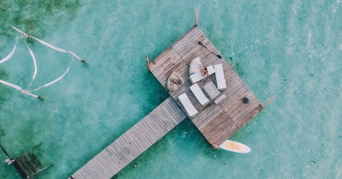 Wooden dock with lounge chairs and a paddleboard on clear turquoise water, a person relaxing at Neek Lagoon Club.
