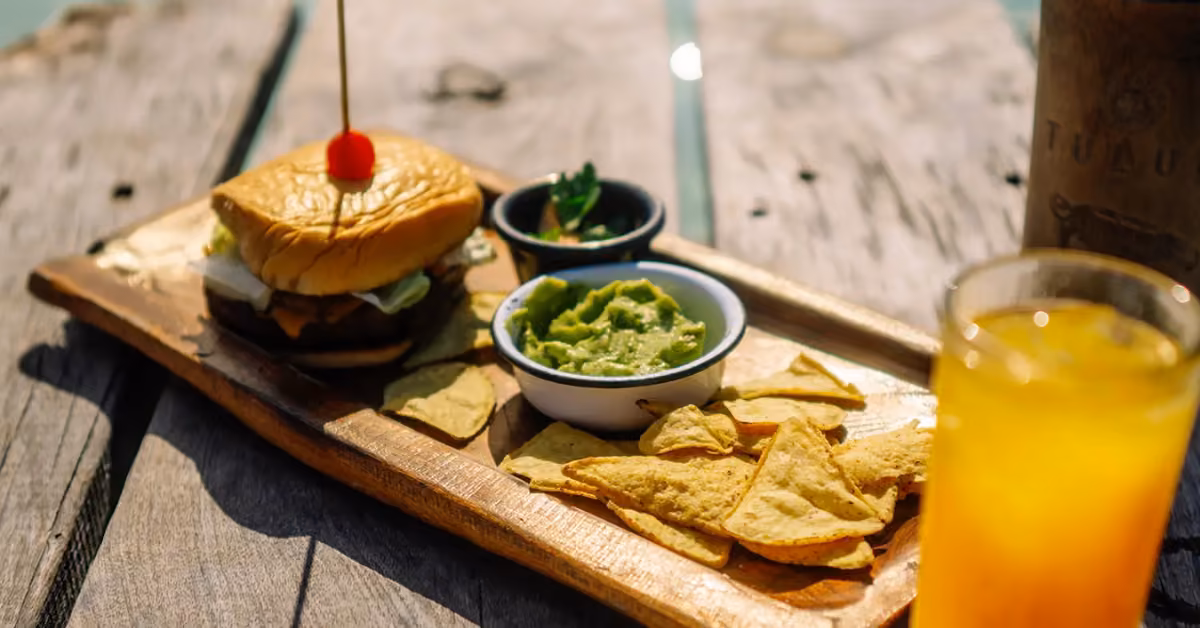 A wooden platter holds a burger with a cherry, guacamole, tortilla chips, salsa, and orange juice on a rustic table.