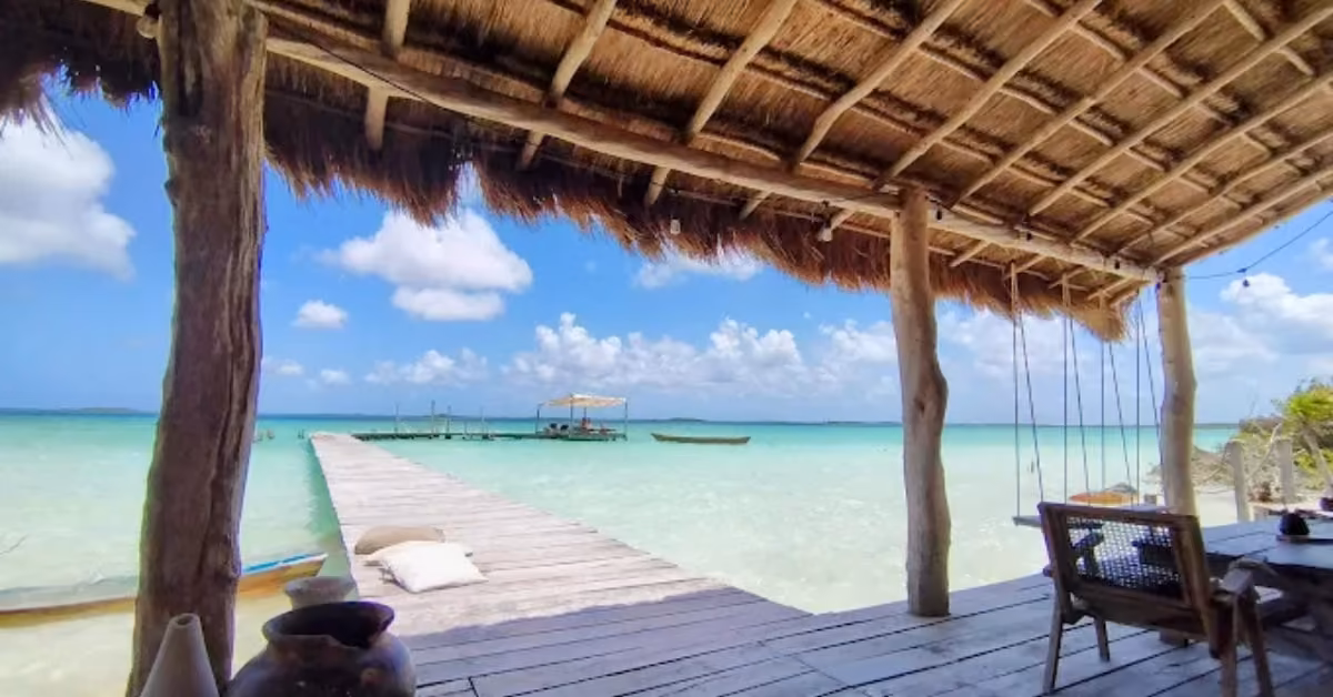 A wooden pier extends over turquoise water from a thatched hut, facing a calm sea and sky. A small boat is docked nearby.
