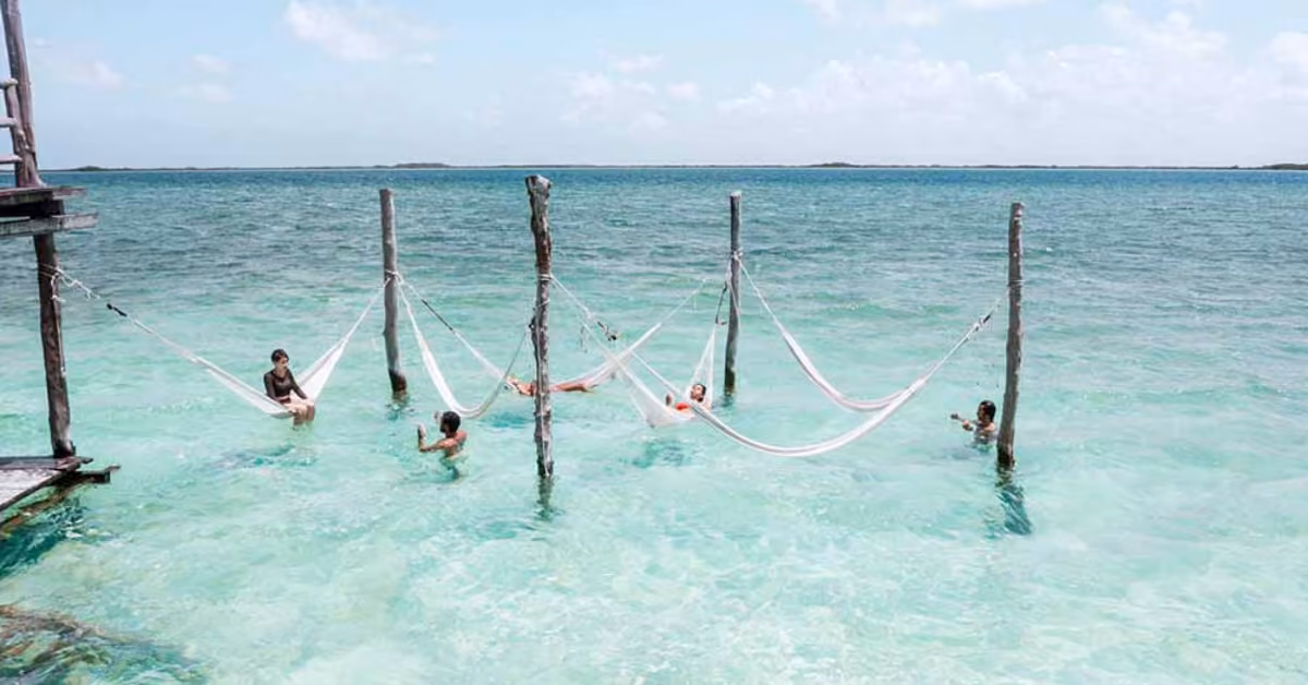People relaxing in hammocks over clear turquoise water at Neek Lagoon Club; a wooden structure and sea under a partly cloudy sky.