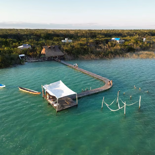 A wooden pier stretches over turquoise water to a thatched hut, with people lounging, swimming, and playing near floating hammocks.