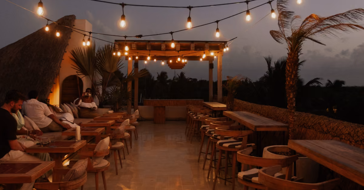 Rooftop bar at sunset with string lights, wooden tables, thatched roof, and palm trees as guests relax in the warm glow.