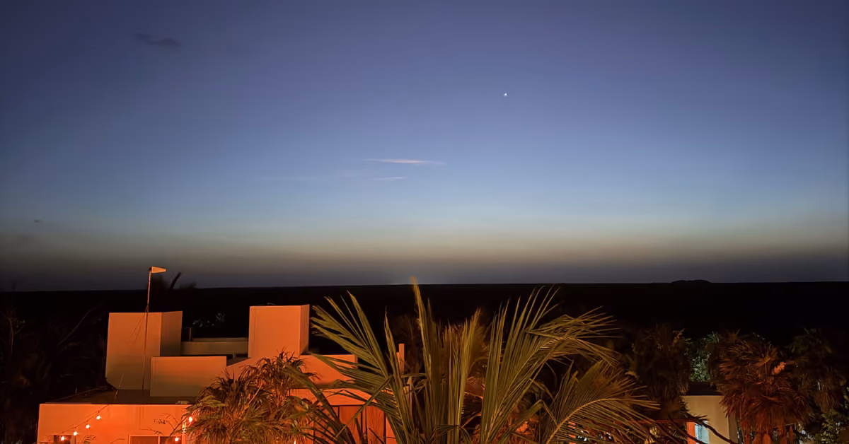 A modern white building and palm trees glow under orange lights at dusk, with a bright star above, seen from the rooftop.