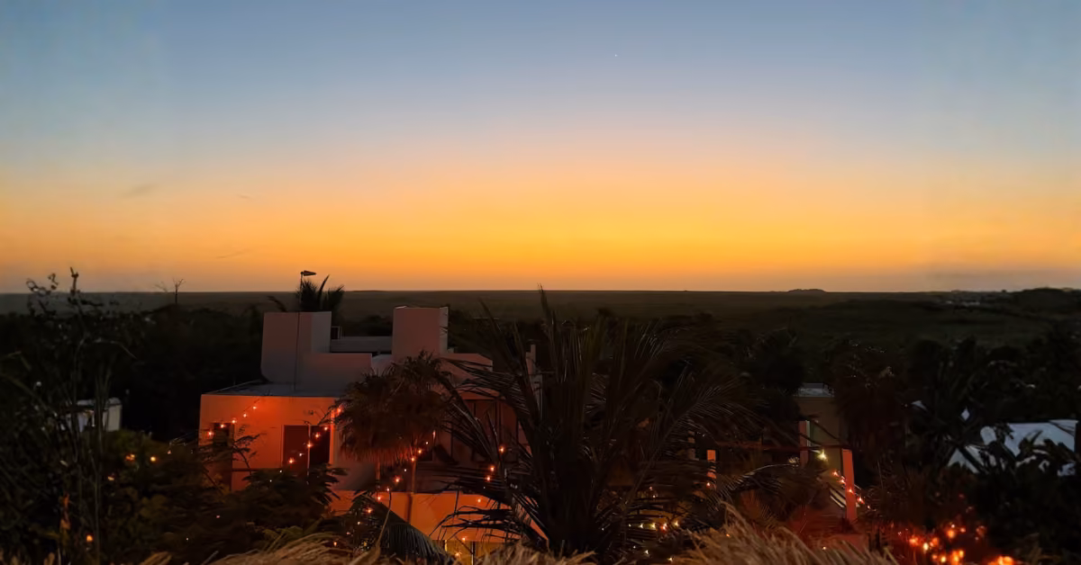 A rooftop with tropical plants and string lights looks out over a wide horizon beneath a vivid orange-blue sunset sky.