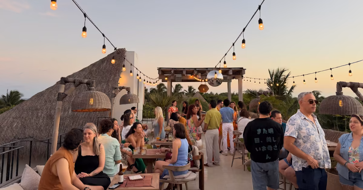 People socializing at Panamera Rooftop at sunset, under string lights, with tropical plants and thatched roofs around cozy tables.