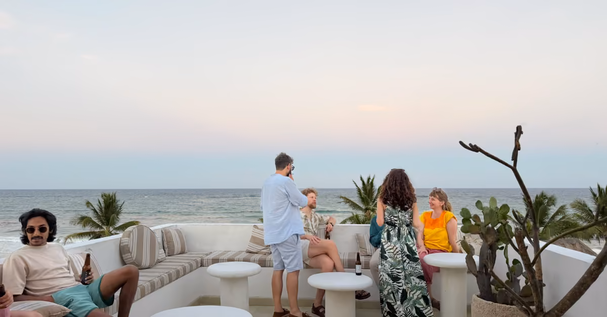 Five people chat on a rooftop lounge with white furniture at sunset, overlooking the ocean and palms. A cactus is on the right.