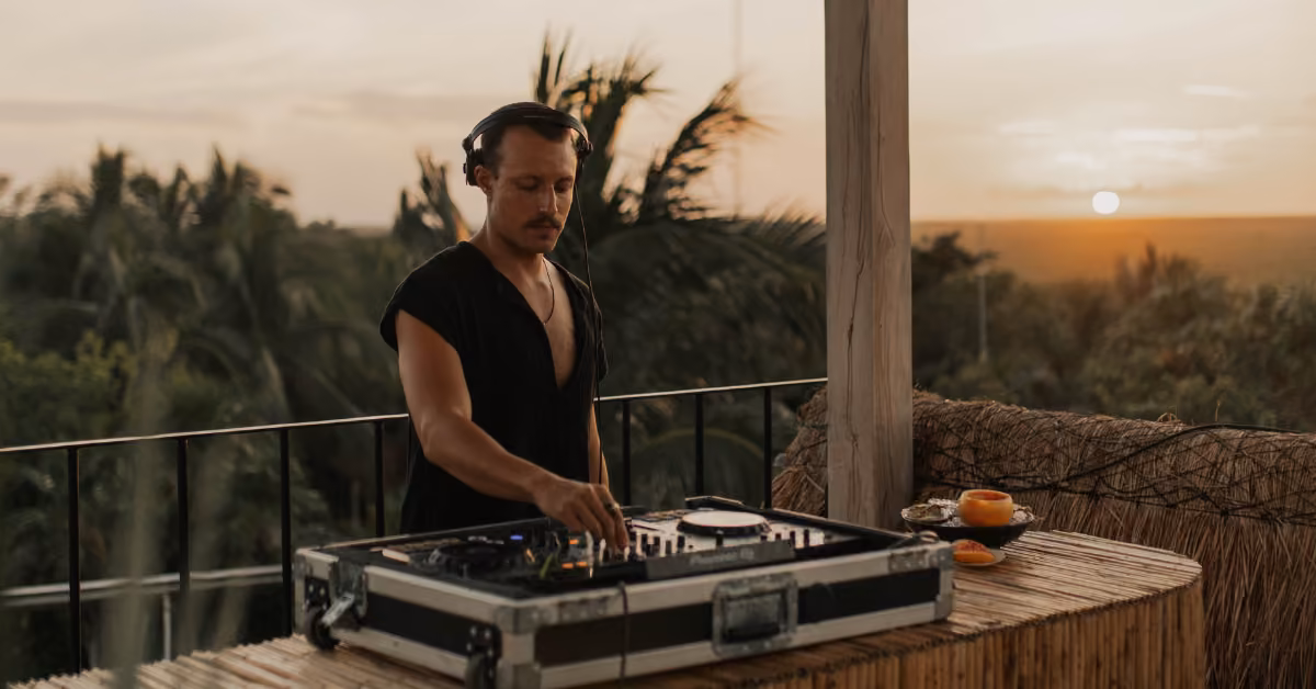 A DJ with headphones mixes music at a rooftop venue lined with palm trees at sunset, fruit arranged on a nearby table.
