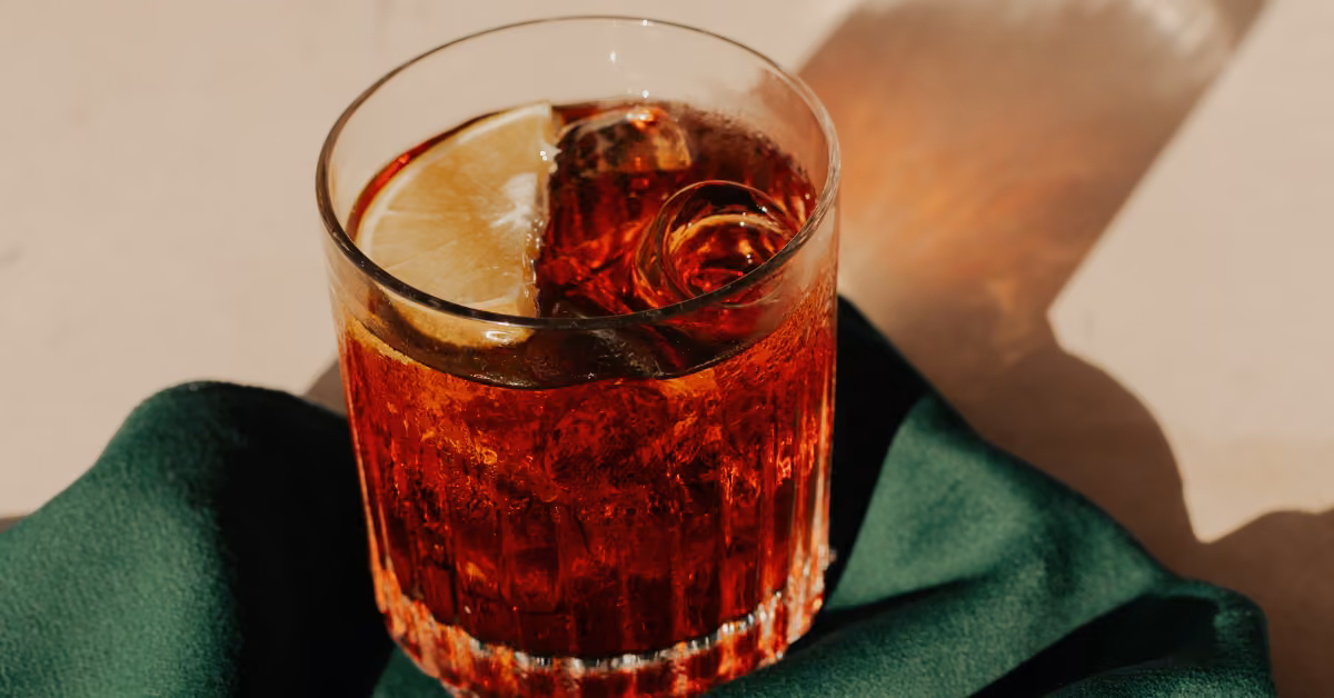 A glass of red cocktail with ice and a lemon slice rests on a green cloth, sunlight casting a shadow on a light surface.