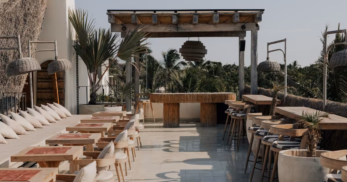 Outdoor bar with wooden tables, barstools, benches, wicker chairs, and plants, overlooking palm trees and a clear sky.