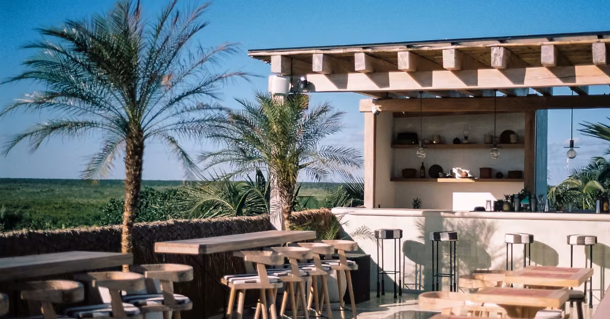 Outdoor bar with wooden tables, palm trees, and a pergola, set against a clear sky and green landscape.