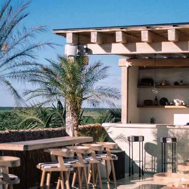 Outdoor café with wooden tables, palm trees, and a bar overlooks a sunlit green landscape under a clear blue sky.