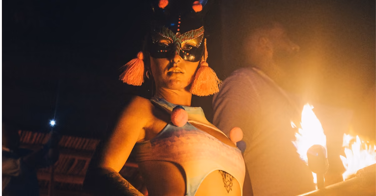 A woman in a vibrant mask and costume stands by a flame at a night club, with a man in the background under dim lights.