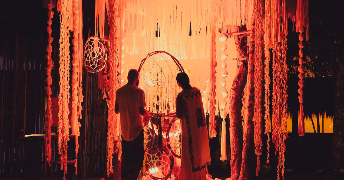 Two people stand before a glowing orange art piece at night, surrounded by dreamcatchers and ribbons in a warm, mystical setting.