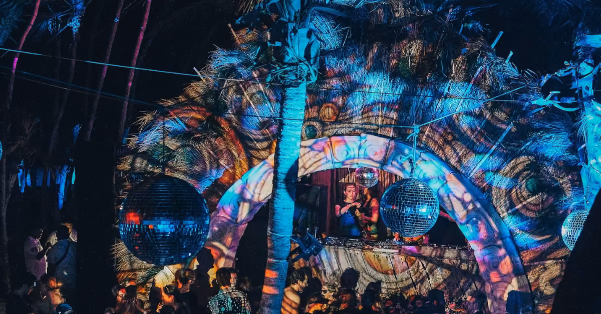 People dance in a lively night club with colorful lights, palm leaves, disco balls, and festive tropical decor on stage.