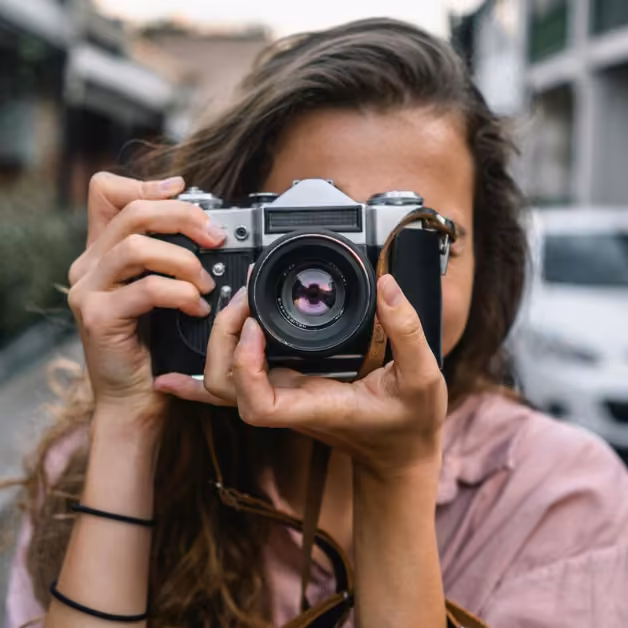 A person with long hair in a light pink shirt holds a vintage camera to their face, standing on a street with a blurred background.